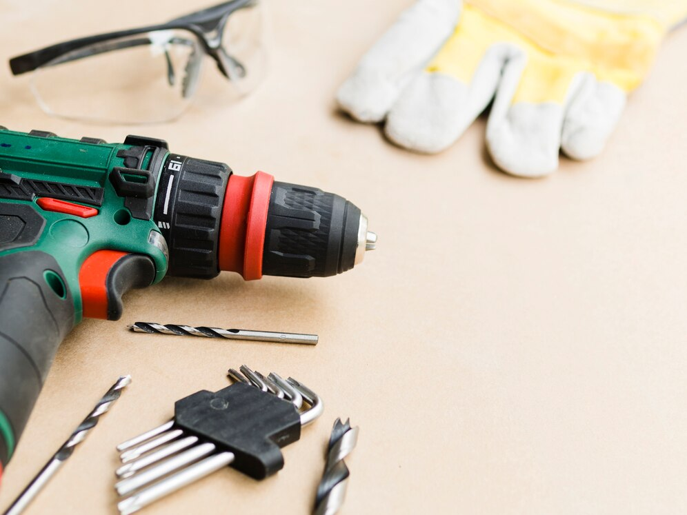 a set of tools laid out on a light-colored surface. In the foreground, there is a green and black power drill with a red and black chuck, accompanied by several drill bits of varying sizes and a hex key set. In the background, a pair of protective safety goggles and yellow work gloves are visible, emphasizing safety precautions