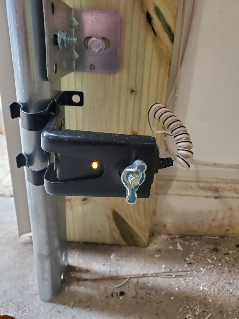 a garage door sensor system mounted on a wooden frame. The sensor housing is black and has a visible indicator light illuminated in yellow, suggesting it is powered on. A coiled wire connects the sensor to the system, with a metal bracket securing it to a vertical support